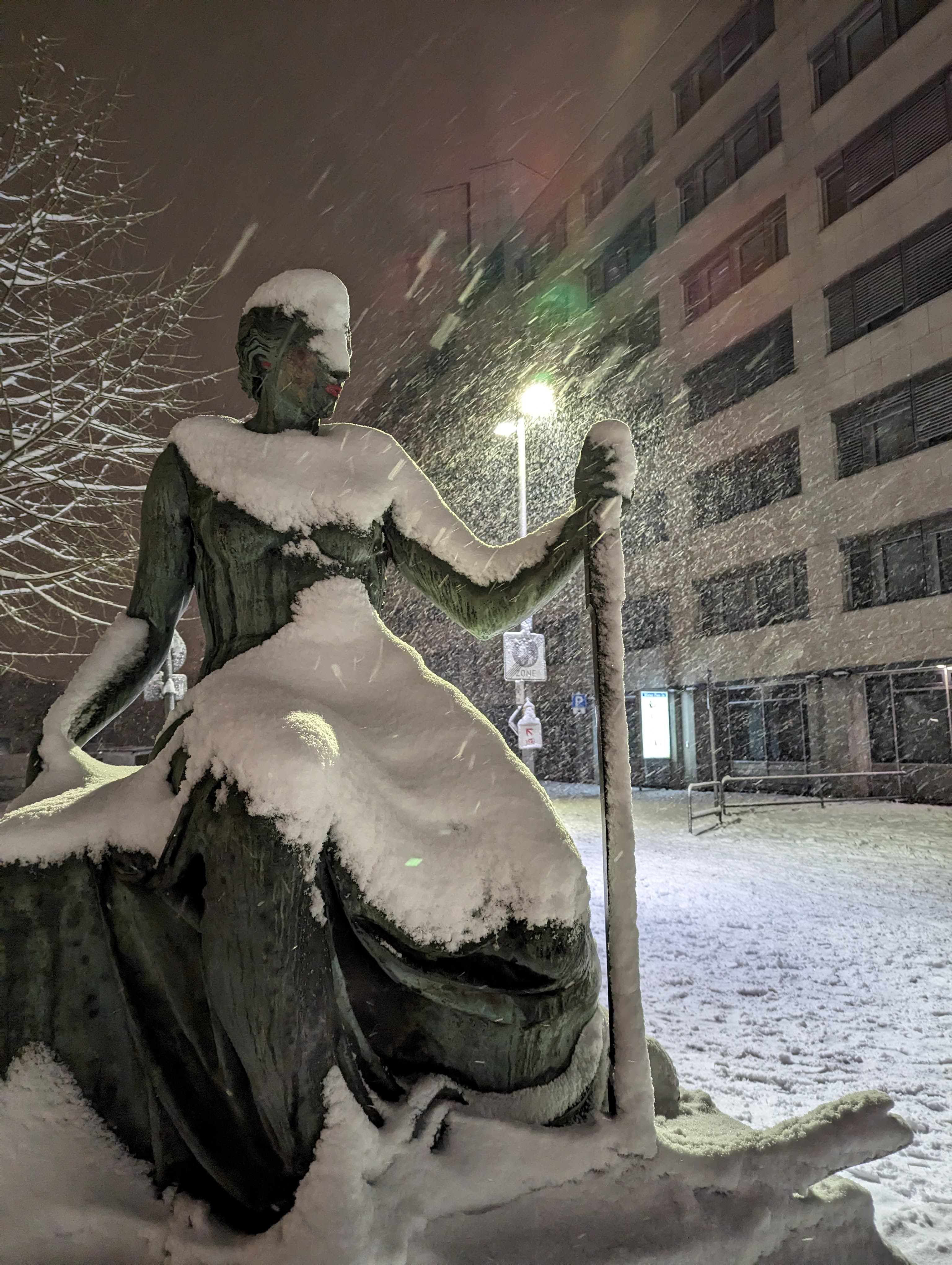 A statue of a women, partially covered in snow, lit from behind by a street lantern under heavy snowfall.